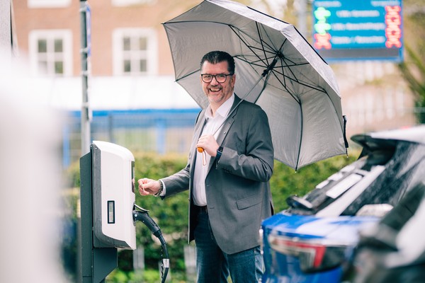 Michael Baune steht mit seinem Elektroauto an der E-Ladesäule und hält eine Chipkarte an diese. Außerdem hat er einen Regenschirm in der Hand.