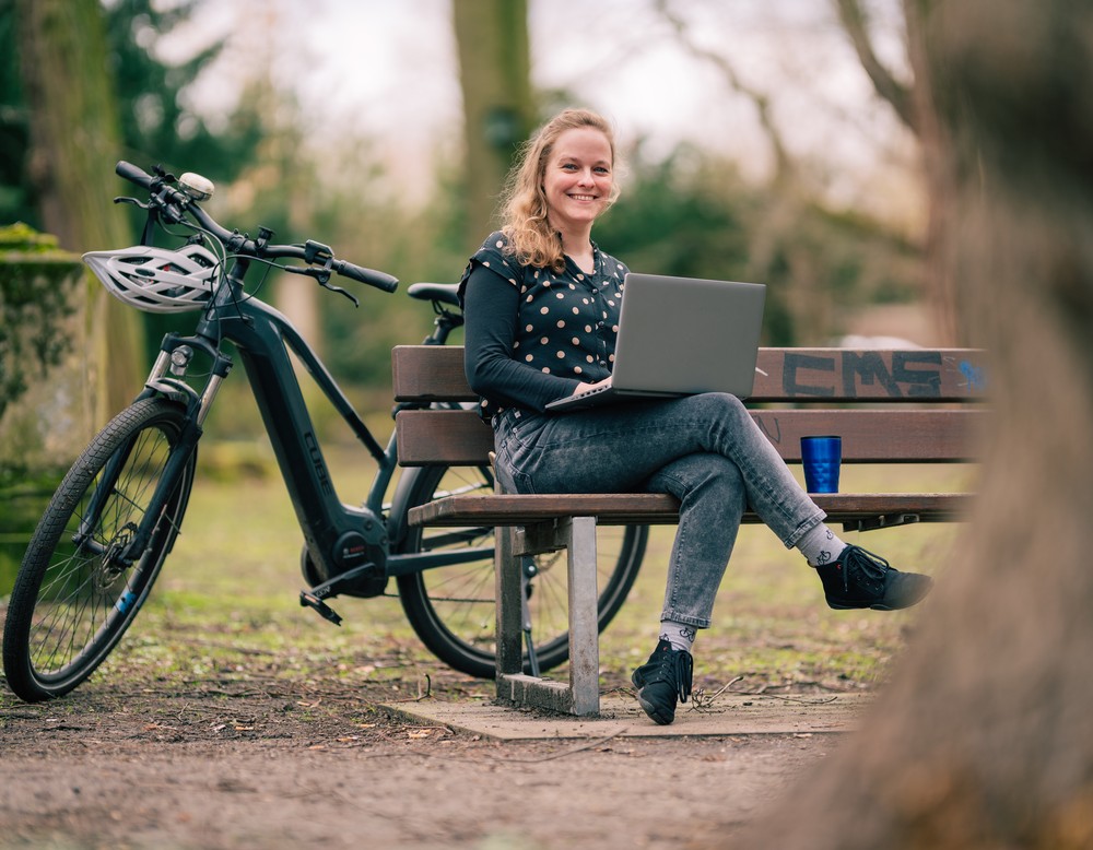 Janina Enning sitzt seitlich auf einer Bank, hinter ihr steht ein Fahrrad und sie hat einen Laptop auf den Knien.