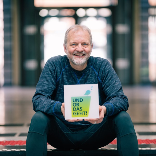 Stefan Rütsch sitzt auf der Treppe und hält die Wort-Bildmarke "Und ob das geht!".