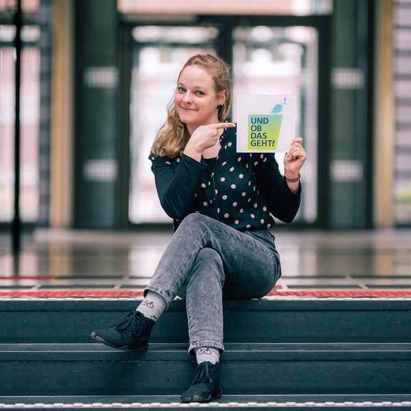 Janina Enning sitzt auf der Treppe und zeigt auf die Wort-Bildmarke "Und ob das geht!".