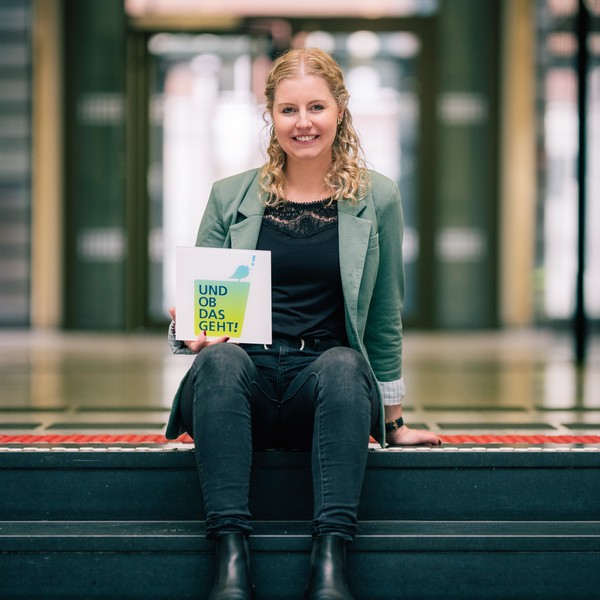 Janine Lammers sitzt auf der Treppe, mit einer Hand abgestützt. Mit der anderen Hand hält sie die Wort-Bildmarke "Und ob das geht!".
