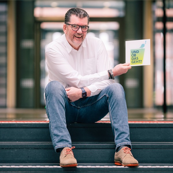 Michael Baune sitzt auf der Treppe und hält die Wort-Bildmarke "Und ob das geht!" seitlich in der Hand.