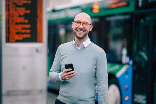 Felix Sewald steht mit seinem Handy in der Hand vor einer Bushaltestelle, hinter ihm ein Bus.